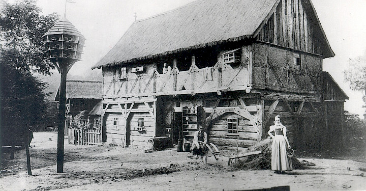 Carnoběła historiska fotografija wjelikego pachanego domu ze słomjanym kšywom. Pśed twarjenim stoje někotare wósoby, mjazy nimi žeńska w serbskej drastwje.