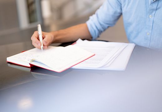 Person in a blue shirt writes with a white pen in an open notebook on a table with other papers