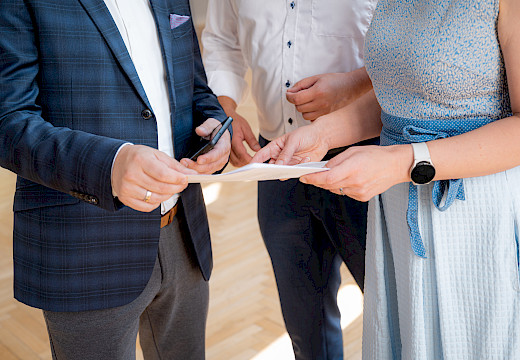 Drei Personen in Businesskleidung übergeben sich ein Blatt Papier, eine Person hält ein Smartphone
