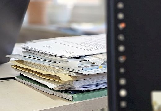 Ein Stapel verschiedener Dokumente und Ordner liegt auf einem Schreibtisch neben einem Computerbildschirm.