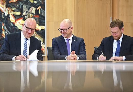 Tśi muske we woblakach sejźe za jadnym blidom, dwa z nich pódpisaju dokumenty, srjejźny muski źaržy ruce złožonej