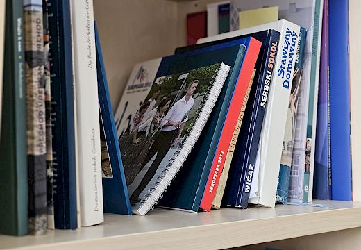 Regal z někotarymi knigłami a spiralnym zešywkom, na kótaremž jo foto dweju wósobowu wiźeś