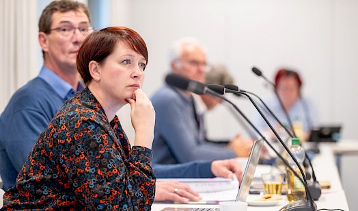 Někotare wósoby sejźe za konferencnym blidom z mikrofonami a laptopami, prědku sejźi žeńska, kenž zepěra brodu na ruku