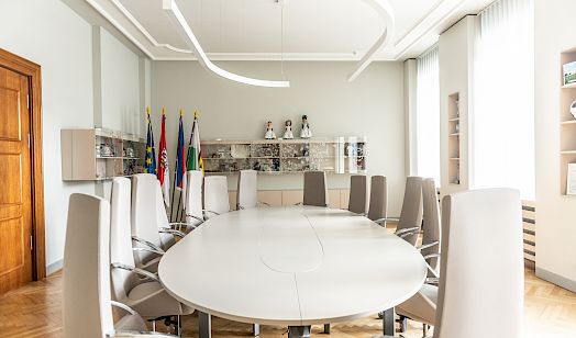 Moderna konferencna rumnosć z owalnym běłym blidom, wusokimi běłymi stołami a někotarymi chórgojami w rožku