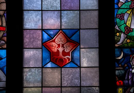 Wokno w Serbskim domje z čerwjenym logom Domowiny z třomi lipowymi łopješkami, kotrež ze zdónka z wosom korjenjemi wuchadźeja, wobdate wot kwadratiskich škleńčanych platow we wšelakich šěrych barbach.