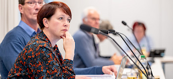 Fenster im Haus der Sorben mit einem roten Domowina-Logo, das drei Lindenblätter zeigt, die aus einem Stamm mit acht Wurzeln hervorsprießen, umgeben von quadratischen Glasfeldern in verschiedenen Grautönen.