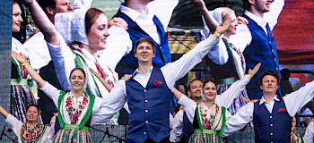 eine Frau und ein Mann geben ein Konzert in einem rustikalen Saal mit Holzbalkendecke vor einem Publikum, das auf Stühlen an Tischen sitzt.