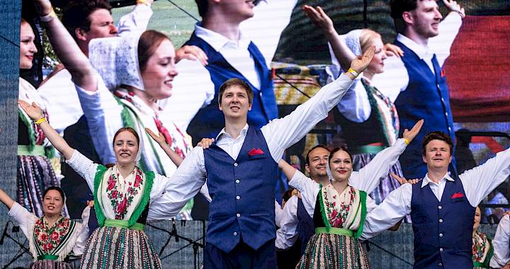 Žeńska a muski koncertěrujotej w rustikalnem zalu z drjewjanym grědowym wjerchom pśed publikumom, kenž na stołach za blidami sejźi.