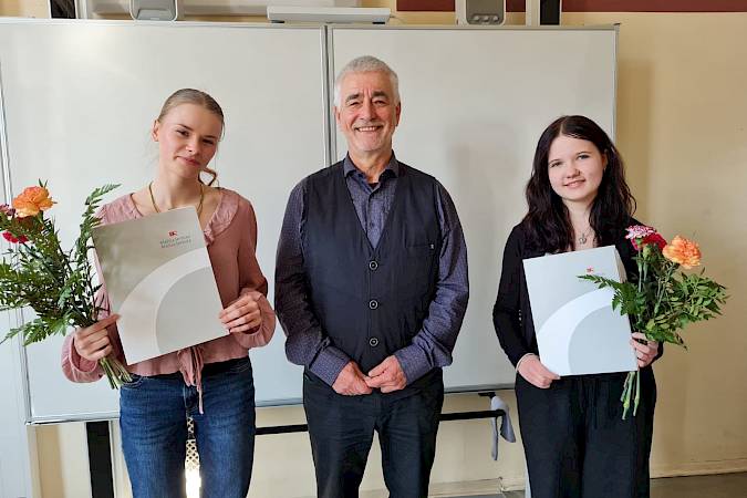 Na dnju zakitowanja semesterskich dźěłow na Delnjoserbskim gymnaziju dnja 19. měrca 2026 spožci předsyda Maśicy Serbskeje Pětš Šurman šulerce Stelli Kliemojc (nalěwo) Myto Bogumiła Šwjele a Luciji Lojchenojc (naprawo) Spěchowanske myto Bogumiła Šwjele.