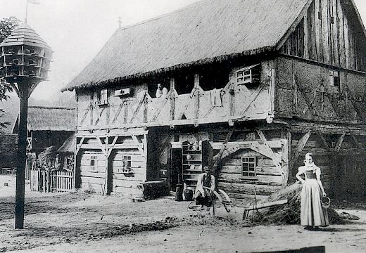 Čornoběła historiska fotografija wulkeho tykowaneho domu ze słomjanskej třěchu. Před domom steja wjacore wosoby, mjez nimi žona w serbskej drasće.