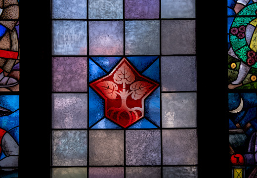 Wokno w Serbskim domje z čerwjenym logom Domowiny z třomi lipowymi łopješkami, kotrež ze zdónka z wosom korjenjemi wuchadźeja, wobdate wot kwadratiskich škleńčanych platow we wšelakich šěrych barbach.