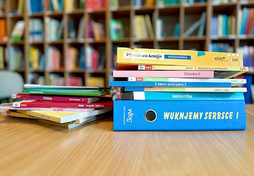 Gestapelte sorbische Schulbücher mit bunten Einbänden auf einem Holztisch vor einem Bücherregal