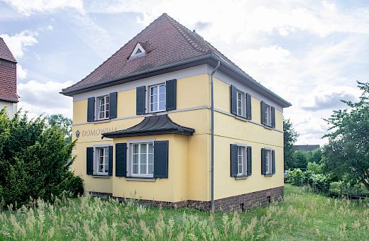 Žołty dwójoschójźeńkowy Domowinski dom we Wórjejcach ze śamnymi woklapnicami a cerwjenym swislowym kšywom, wobdany wót wusokeje tšawy a bomow