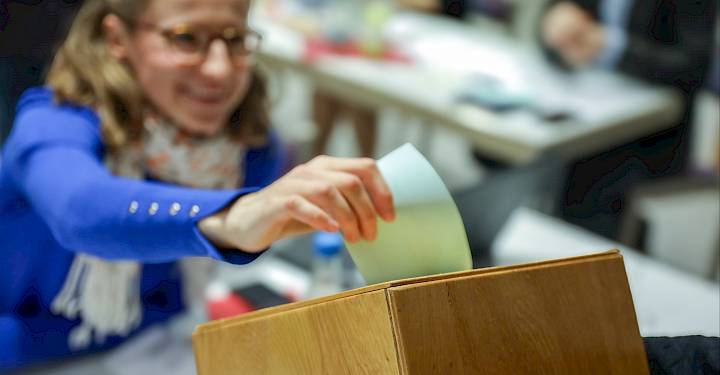 Frau mit blauem Oberteil wirft Zettel in eine hölzerne Wahlurne