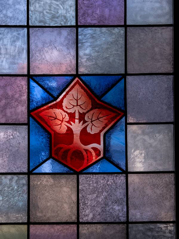 Fenster im Haus der Sorben mit einem roten Domowina-Logo, das drei Lindenblätter zeigt, die aus einem Stamm mit acht Wurzeln hervorsprießen, umgeben von quadratischen Glasfeldern in verschiedenen Grautönen.