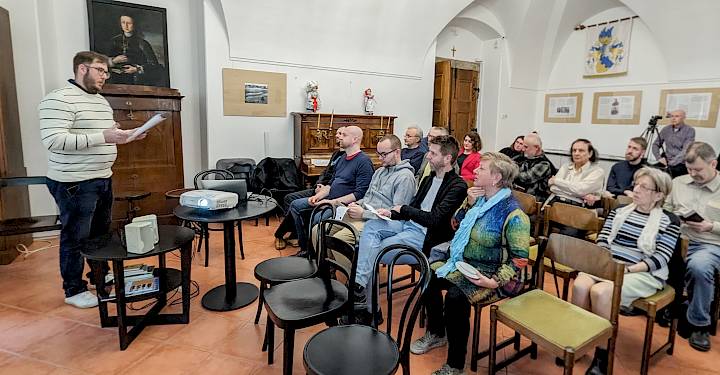 Mann mit gestreiftem Pullover hält Vortrag vor sitzendem Publikum in einem Raum mit Klavier und Gemälde