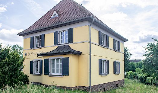 Gelbes zweistöckiges Domowina-Haus in Hoyerswerda mit dunklen Fensterläden und rotem Satteldach umgeben von hohem Gras und Bäumen